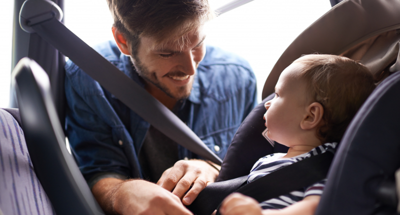 Et barn sidder i en autostol, mens barnets far smiler til barnet.
